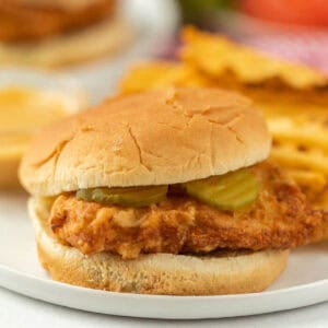 A crispy fried chicken sandwich reminiscent of a copycat Chick-fil-A recipe is paired with pickles on a soft bun, elegantly served on a white plate. In the background, slightly out of focus, are golden chips adding to the inviting scene.