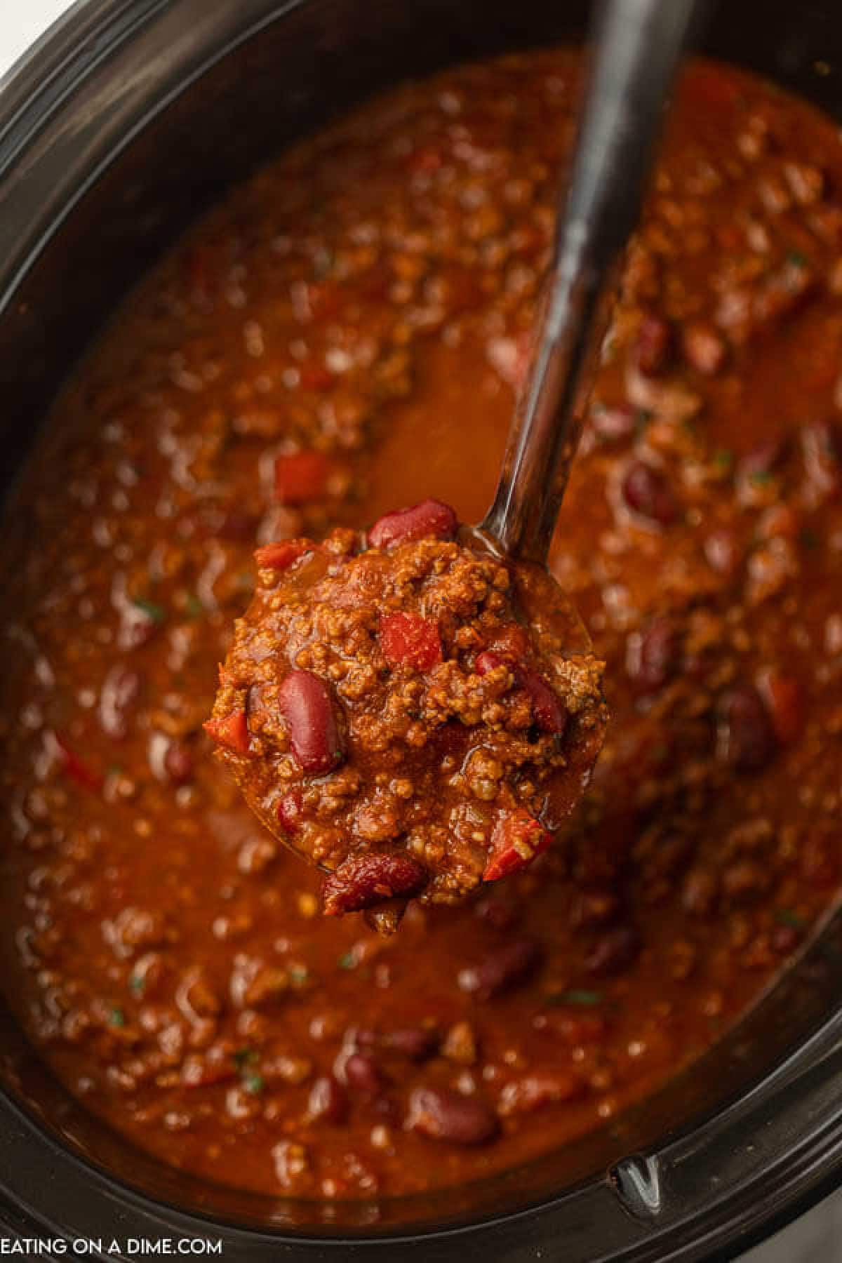 A ladle lifting a serving of Crockpot Cowboy Chili from a slow cooker. The chili is rich and chunky, filled with ground meat, red kidney beans, and diced tomatoes, all in a thick, savory sauce.