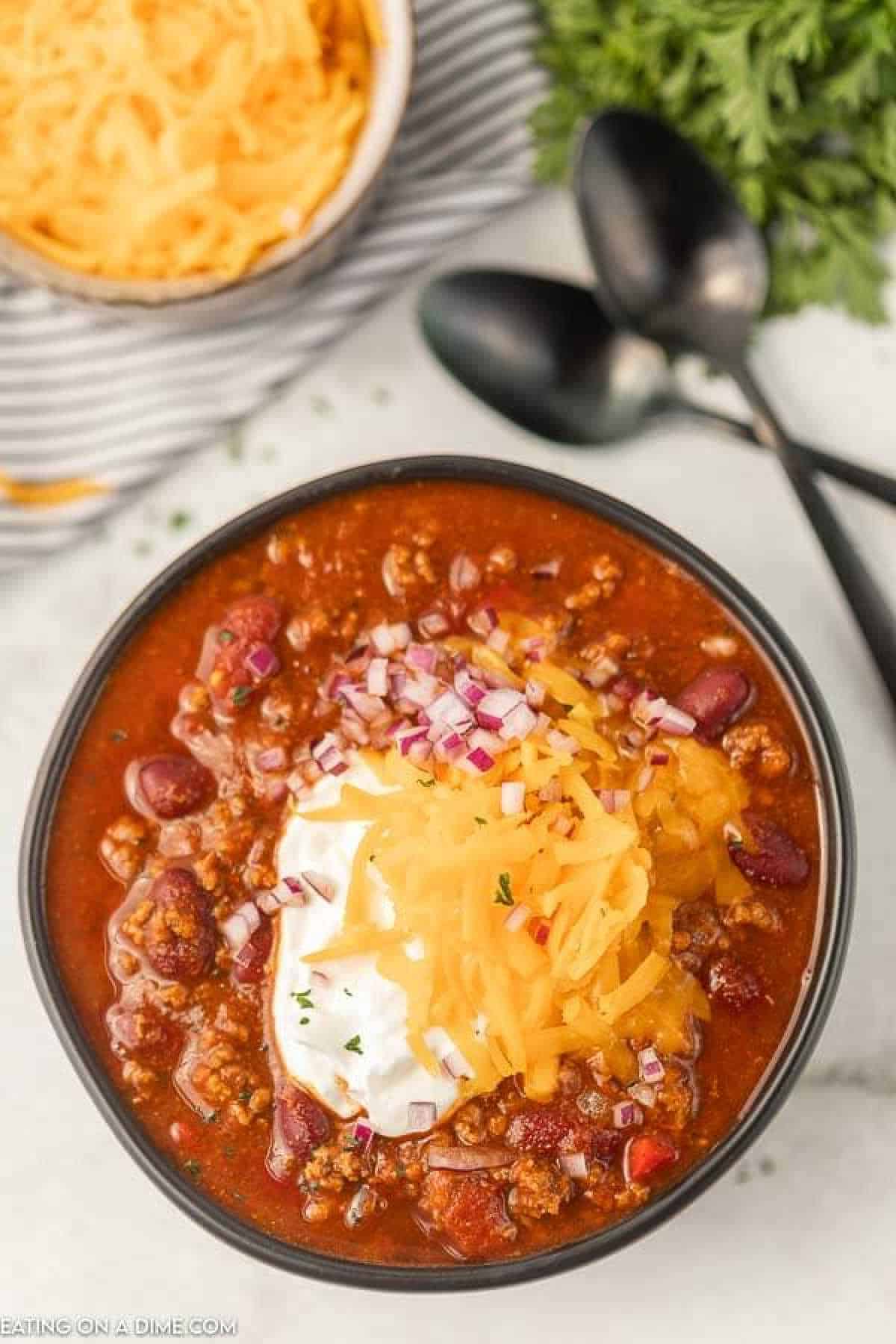 A bowl of Crockpot Cowboy Chili topped with sour cream, shredded cheese, and chopped red onions. The hearty mix features kidney beans and ground meat. In the background, there’s a bowl of shredded cheese, three black spoons, and fresh green herbs.