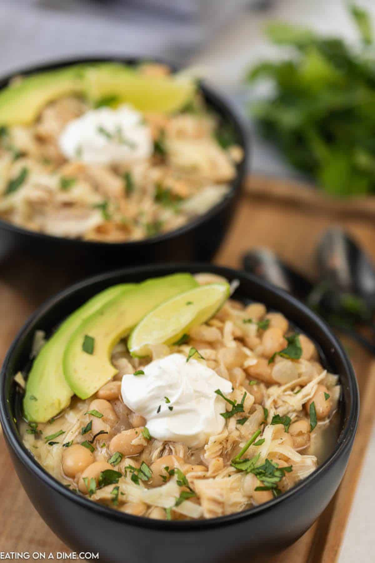  Close up image of two black bowls of white chicken chili with avocado on a board. 