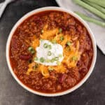 A bowl of savory Instant Pot chili is topped with shredded cheese, sour cream, and chopped green onions. This hearty dish contains ground meat and kidney beans, resting on a dark surface with fresh green onions in the background.