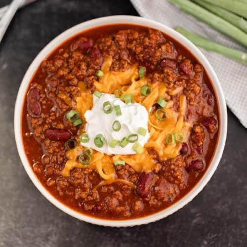 A bowl of savory Instant Pot chili is topped with shredded cheese, sour cream, and chopped green onions. This hearty dish contains ground meat and kidney beans, resting on a dark surface with fresh green onions in the background.