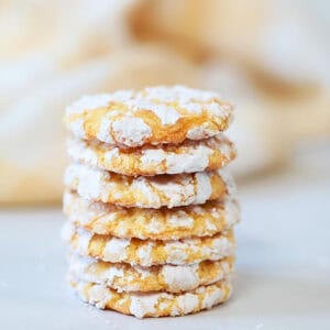 A stack of seven lemon crinkle cookies with a light yellow hue, each dusted with powdered sugar, is placed on a smooth white surface. The soft focus background is a creamy beige, adding warmth to the image.