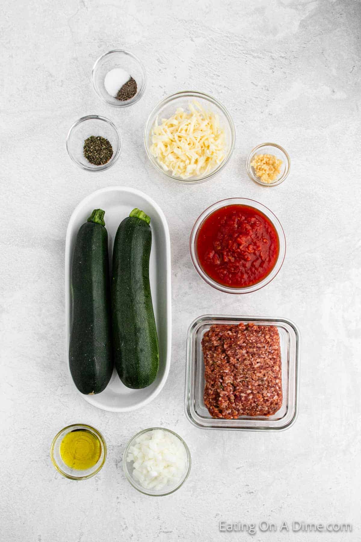 Ingredients for this stuffed zucchini boats recipe: two whole zucchinis, ground meat, marinara sauce, shredded cheese, minced garlic, chopped onions, olive oil, dried basil, salt, and pepper—neatly arranged on a light-colored countertop.