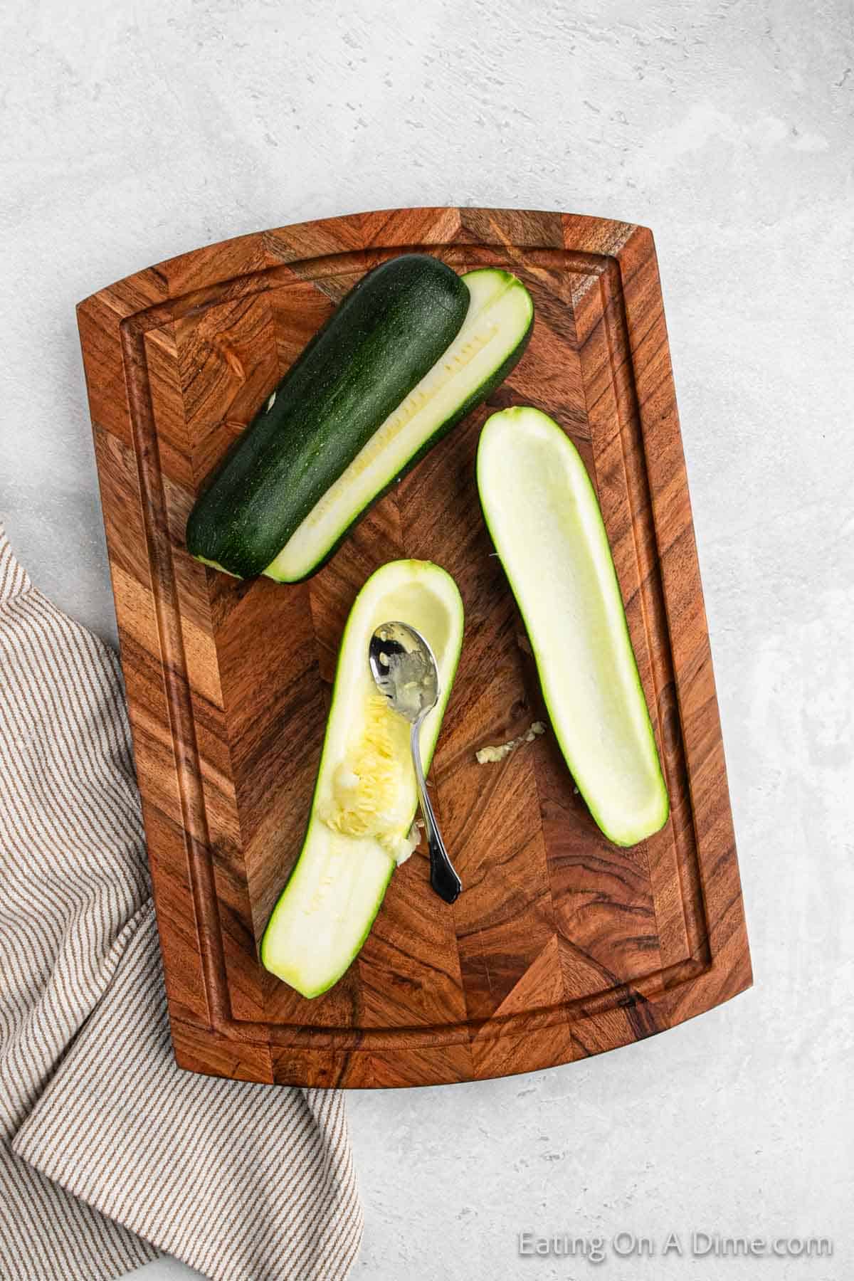 A wooden cutting board displays two zucchini, one of which is halved and partially hollowed out with a spoon, prepped for a stuffed zucchini boats recipe. A striped kitchen towel is partially visible on the left side of the board, resting on a light gray surface.