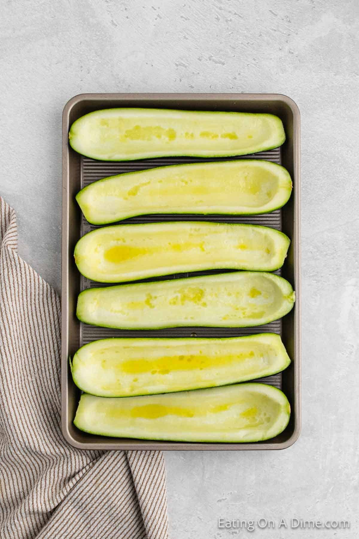 A baking tray with six halved and scooped-out zucchini, brushed with oil or butter, arranged neatly in two rows of three. Perfect for a stuffed zucchini boats recipe, the tray is placed on a light gray surface next to a striped kitchen towel.