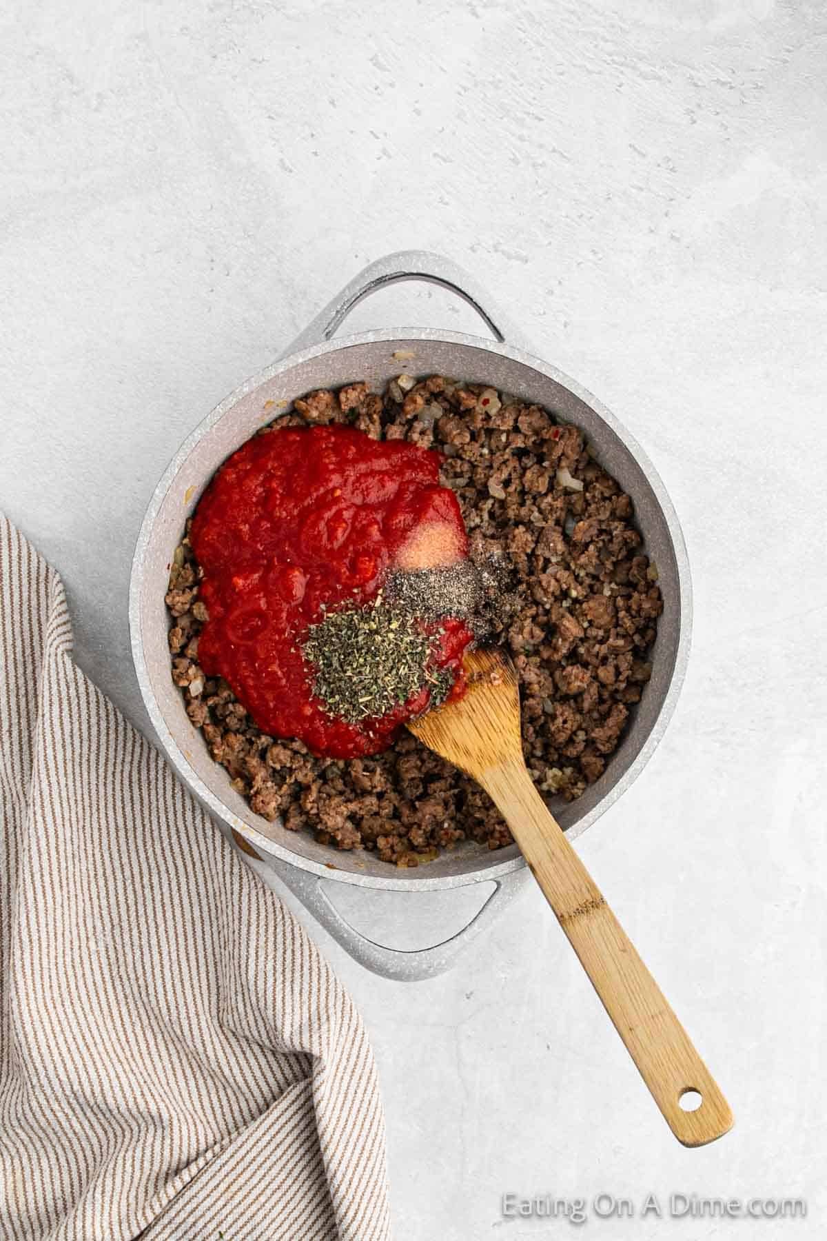A pot filled with cooked ground beef topped with tomato sauce and sprinkled with herbs. A wooden spoon rests inside the pot, which is placed on a light gray surface next to a beige striped cloth—perfect for your next stuffed zucchini boats recipe.
