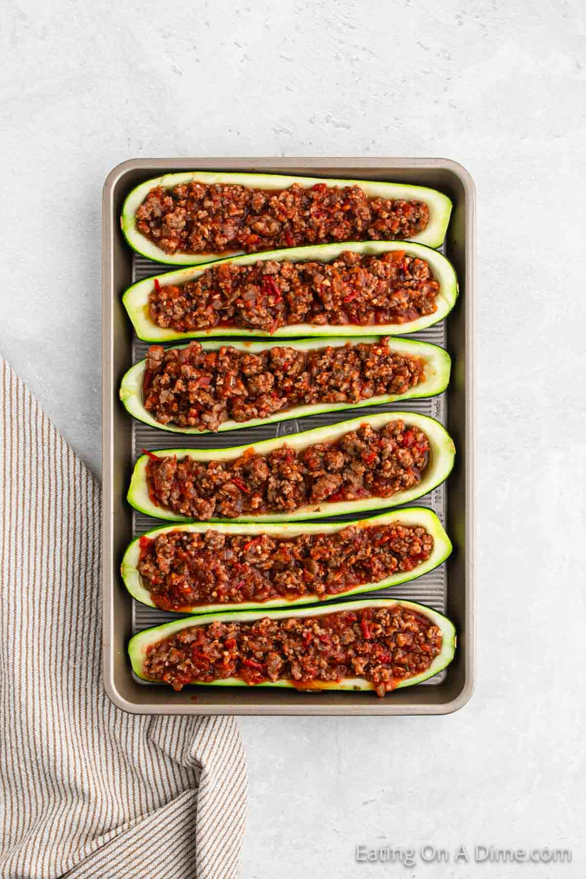 A baking tray with six zucchini halves stuffed with a ground meat and tomato filling, arranged in two rows. The stuffed zucchini boats recipe is displayed on a light-colored surface with a beige and white striped cloth placed next to it.