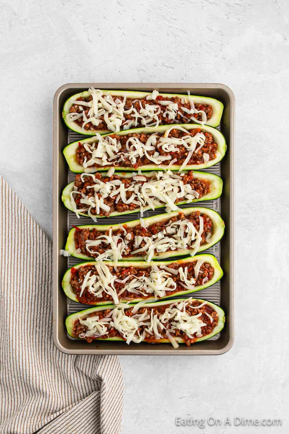 A baking tray filled with six hollowed zucchini halves, stuffed with a mixture of ground meat, sauce, and topped with shredded cheese, ready to be baked. A striped kitchen towel is placed to the left of the tray on a light-colored countertop—a delicious stuffed zucchini boats recipe in the making.