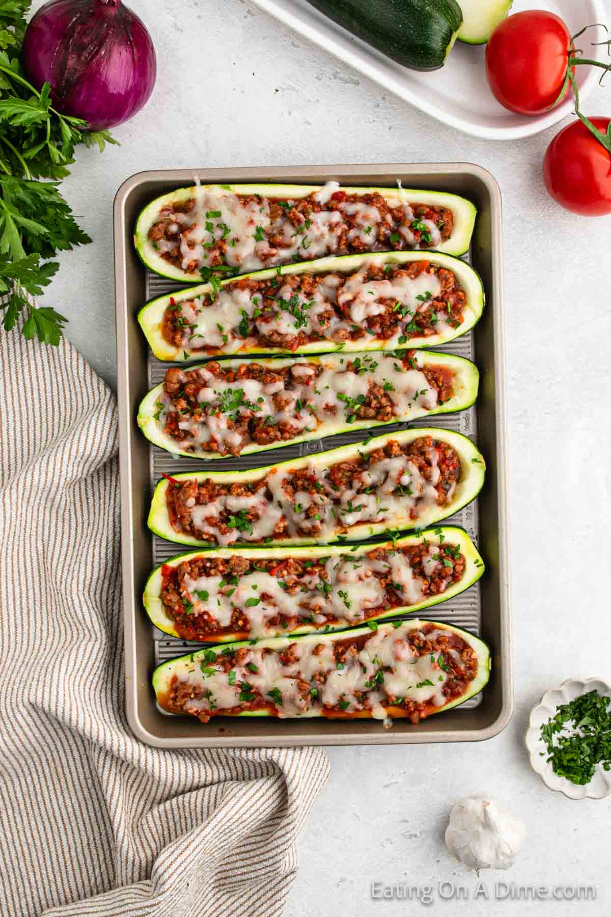 A baking sheet holds halved zucchini boats stuffed with a savory meat and tomato mixture, topped with melted cheese and garnished with parsley. Nearby, there are fresh tomatoes, garlic, a red onion, zucchini, parsley, and a striped kitchen towel—perfect for recreating this stuffed zucchini boats recipe.
