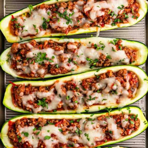 Four stuffed zucchini boats, filled with a mixture of ground meat, diced vegetables, and topped with melted cheese and chopped herbs, arranged on a baking tray. The dish appears colorful and appetizing, showcasing a blend of green, red, and brown ingredients.