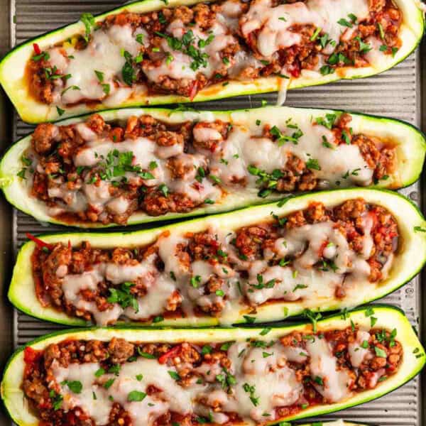 Four stuffed zucchini boats, filled with a mixture of ground meat, diced vegetables, and topped with melted cheese and chopped herbs, arranged on a baking tray. The dish appears colorful and appetizing, showcasing a blend of green, red, and brown ingredients.