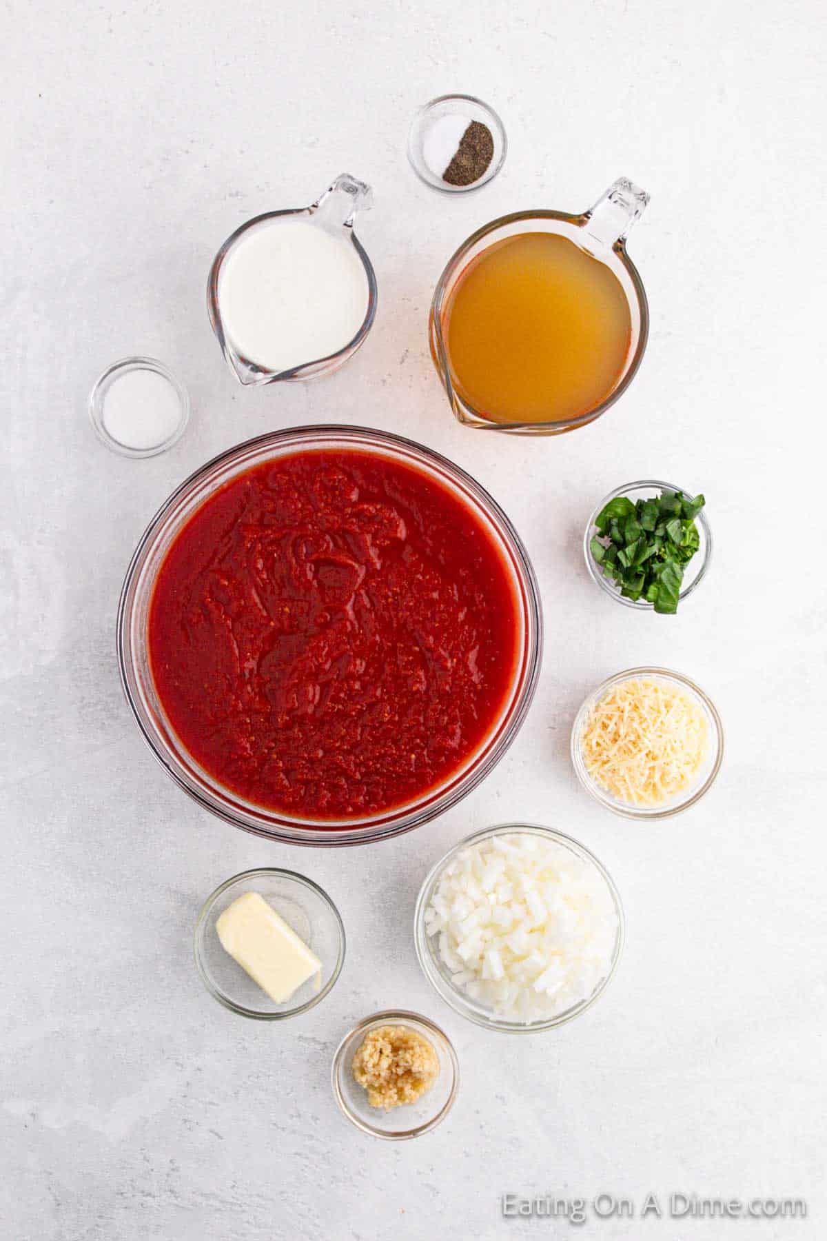 A variety of ingredients for a rich tomato soup recipe are elegantly arranged on a light surface: a bowl of tomato puree, diced onions, grated cheese, butter, minced garlic, fresh basil, milk, broth, and small bowls of salt and pepper.