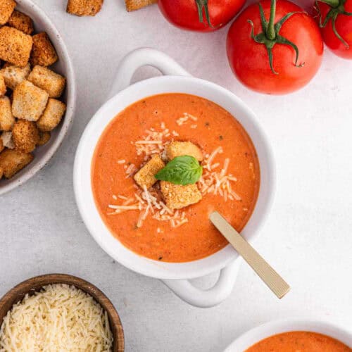 Discover the ultimate tomato soup recipe with a bowl of creamy delight topped with grated cheese, croutons, and a basil leaf. Nestled among fresh vine tomatoes, extra croutons, and a bowl of grated cheese, it’s the perfect comfort on a light surface.