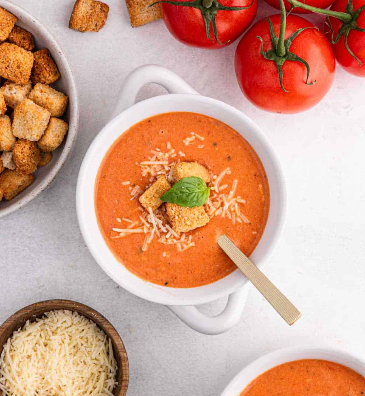 Discover the ultimate tomato soup recipe with a bowl of creamy delight topped with grated cheese, croutons, and a basil leaf. Nestled among fresh vine tomatoes, extra croutons, and a bowl of grated cheese, it’s the perfect comfort on a light surface.