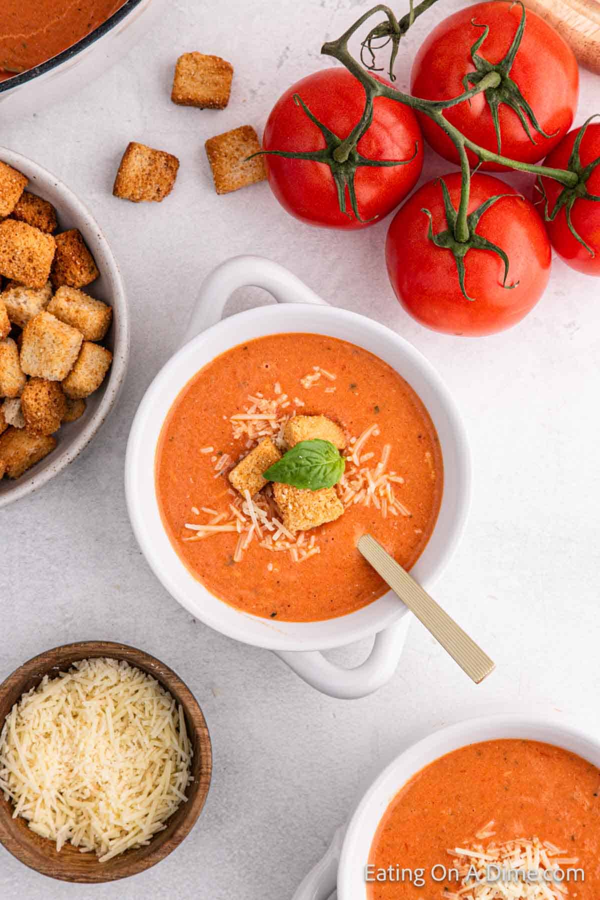 A bowl of delightful tomato soup recipe features croutons, grated cheese, and a basil leaf. Nearby, fresh tomatoes and an extra helping of croutons rest beside a bowl of shredded cheese. A spoon eagerly awaits in the comforting goodness.