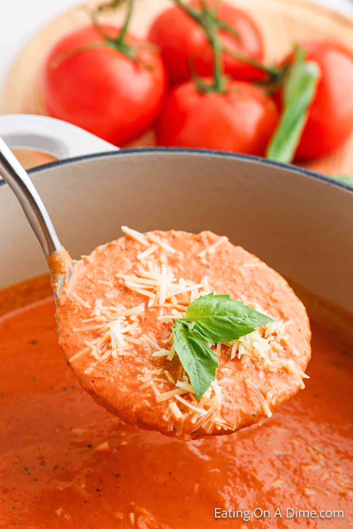 A ladle lifts creamy tomato basil soup from a pot, showcasing a delicious tomato soup recipe topped with grated cheese and fresh basil leaves. In the background, ripe tomatoes and basil rest on a wooden cutting board. The scene is bright and appetizing.