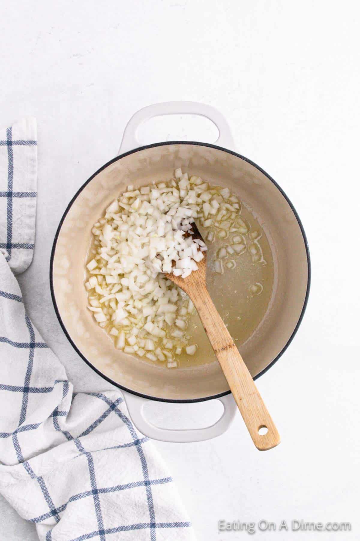 A pot with diced onions being sautéed in oil, the perfect base for a rich tomato soup recipe. A wooden spoon gently stirs the onions while a blue and white checkered kitchen towel peeks from the side.