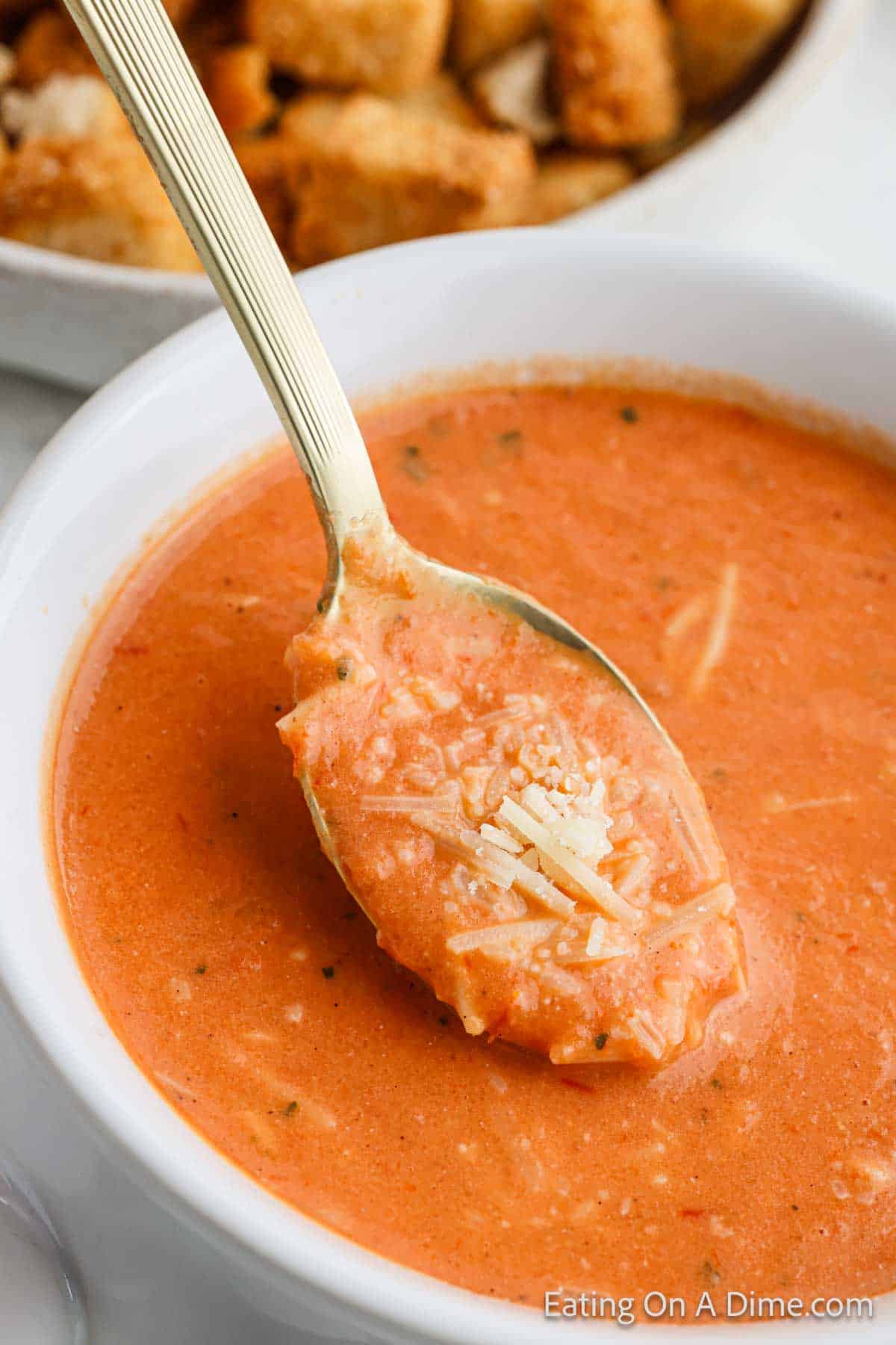 A close-up of a spoonful of creamy tomato parmesan soup being lifted from a white bowl, embodying the perfect tomato soup recipe. The soup is garnished with shredded cheese, while a plate of croutons tempts in the background.