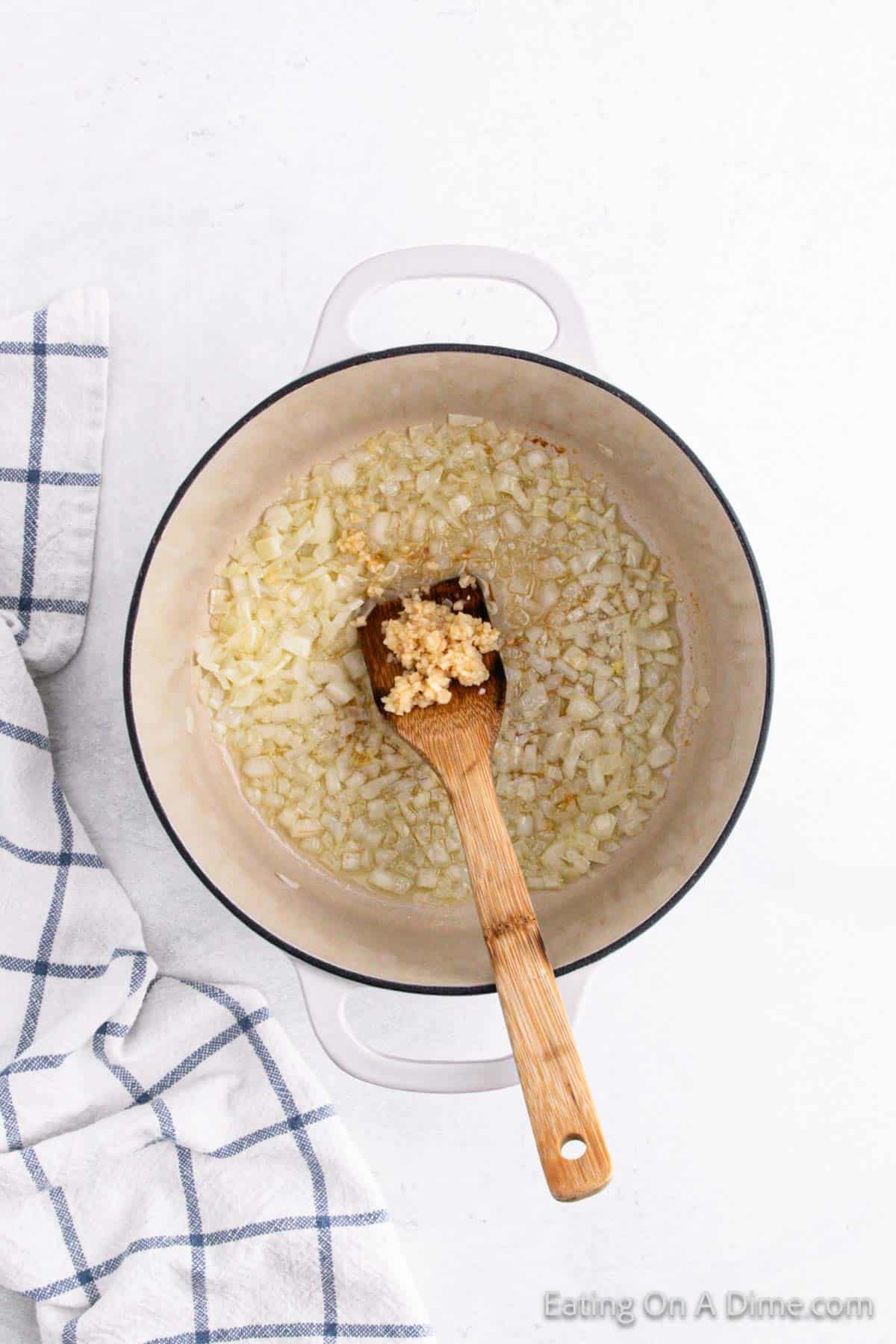 A pot with sautéed onions and minced garlic being stirred with a wooden spoon, forming the fragrant base of a delicious tomato soup recipe. A white and blue checkered cloth is partially visible on the left side, all set on a light-colored surface.