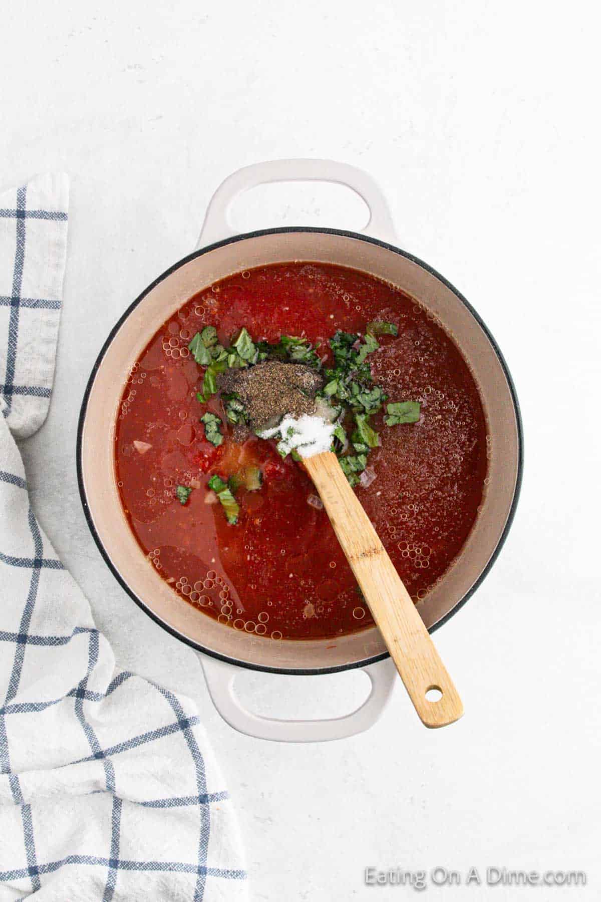 A pot of delicious tomato soup recipe with chopped herbs and seasoning on top, stirred with a wooden spoon. A blue and white checkered cloth is partially visible to the left.