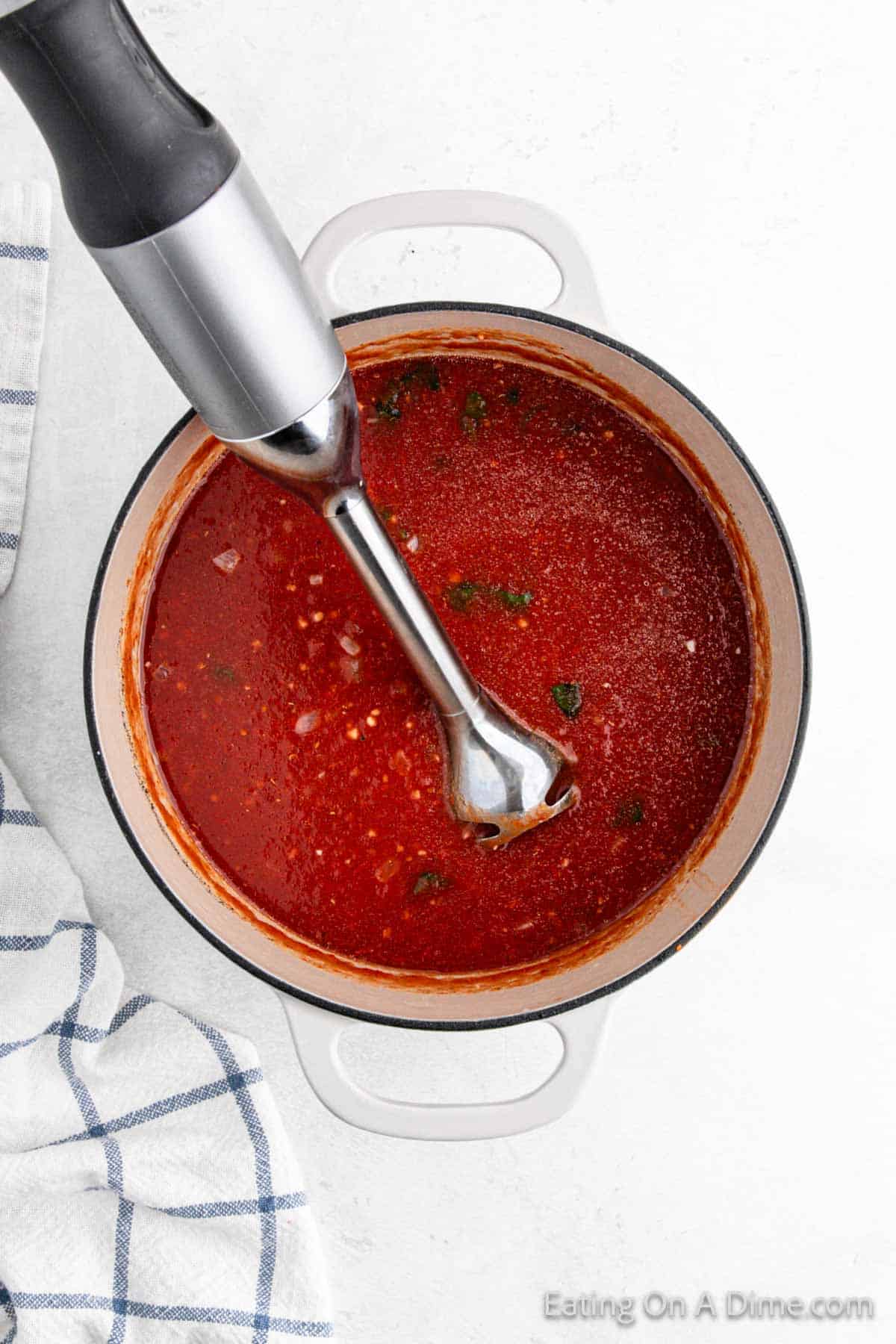 A top view of a pot showcasing a delicious tomato soup recipe being blended with an immersion blender. The pot sits on a white surface, accompanied by a striped kitchen towel.