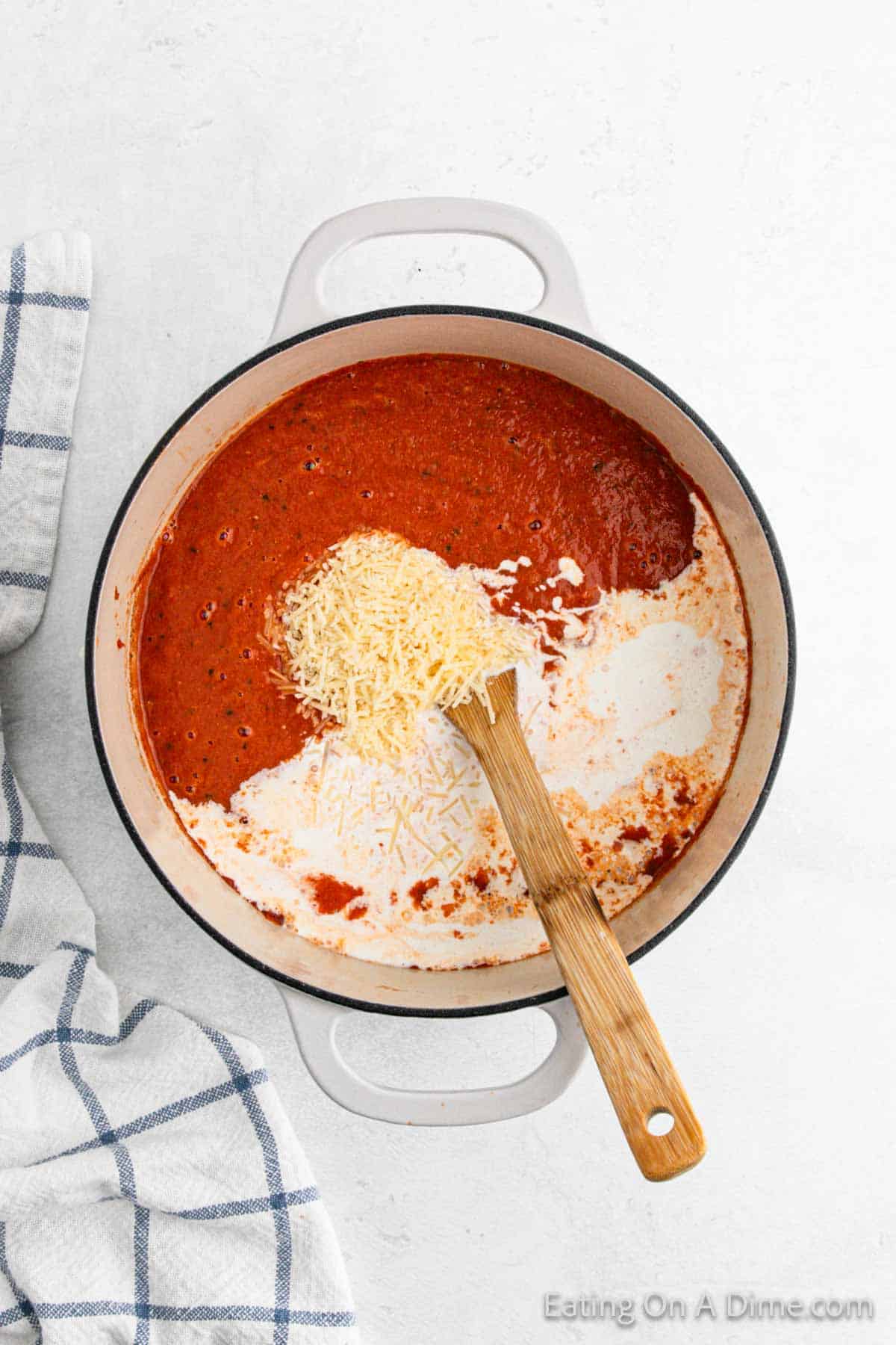 A pot containing a delightful tomato soup recipe swirls with cream and grated cheese as a wooden spoon stirs. Nearby, a blue and white striped cloth rests on the light surface, adding a touch of charm.