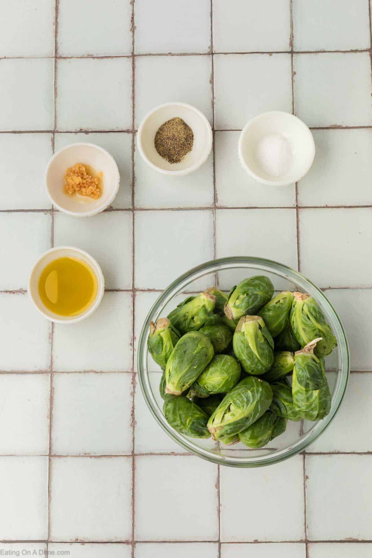 A glass bowl filled with Brussels sprouts is centered on a tiled surface. Surrounding the bowl are small white dishes containing chopped garlic, black pepper, salt, and olive oil—perfect for prepping an air fryer Brussels sprouts recipe. The layout appears to be for a cooking preparation.