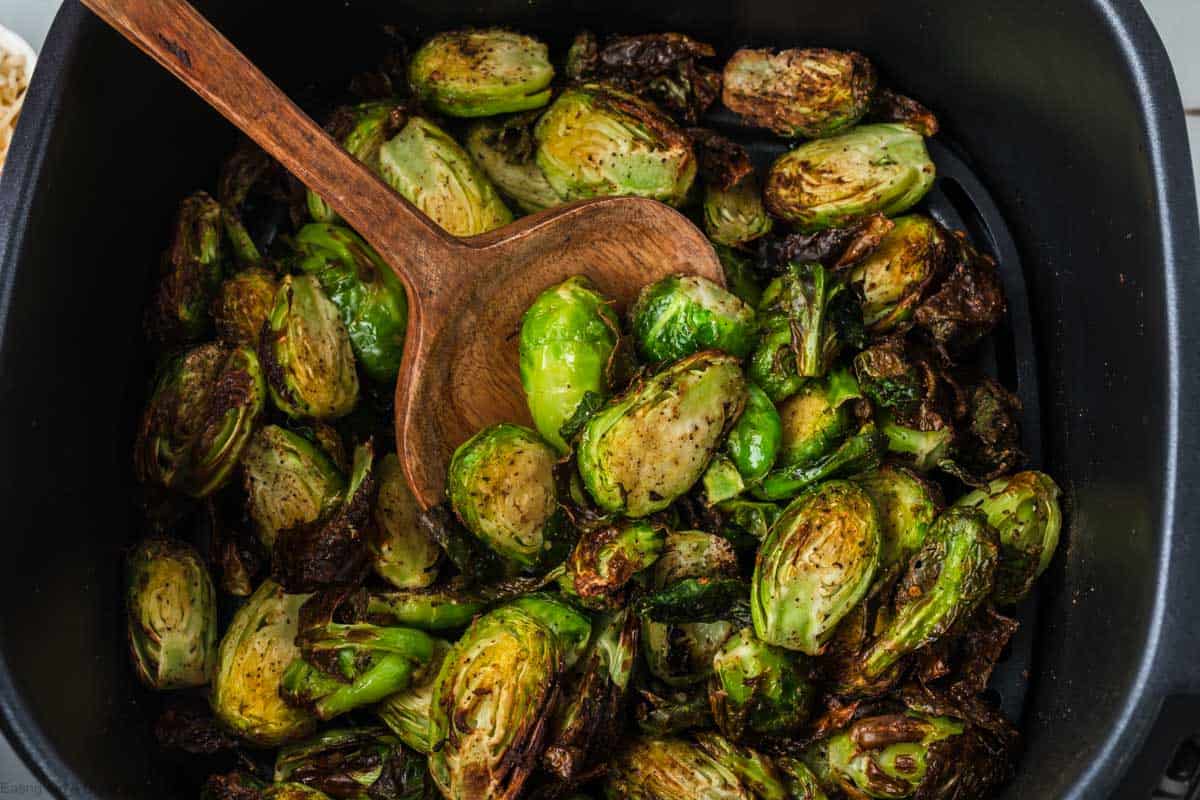 A close-up of roasted Brussels sprouts in a black tray. The sprouts are browned and caramelized, indicating they were likely cooked to a crispy texture using an air fryer. A wooden spoon is nestled among them, suggesting they are ready to be served or stirred.