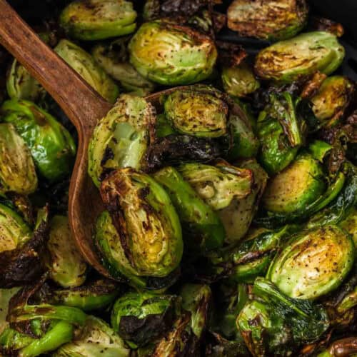 Close-up of roasted Brussels sprouts in a bowl with a wooden spoon scooping some. The Brussels sprouts are browned, slightly charred, and seasoned with visible specks of spices, giving them a crispy and flavorful appearance—a perfect result from following an air fryer Brussels sprouts recipe.