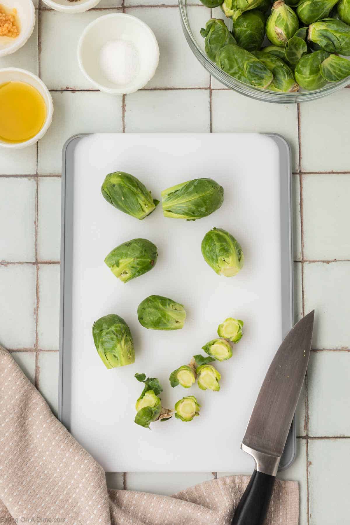 A cutting board with trimmed Brussels sprouts and their stalk ends. A knife lies to the right, with a beige cloth nearby. On the top right, a bowl contains more Brussels sprouts ready for an air fryer Brussel sprouts recipe. Small bowls of seasoning and oil are in the top left corner on a tiled surface.