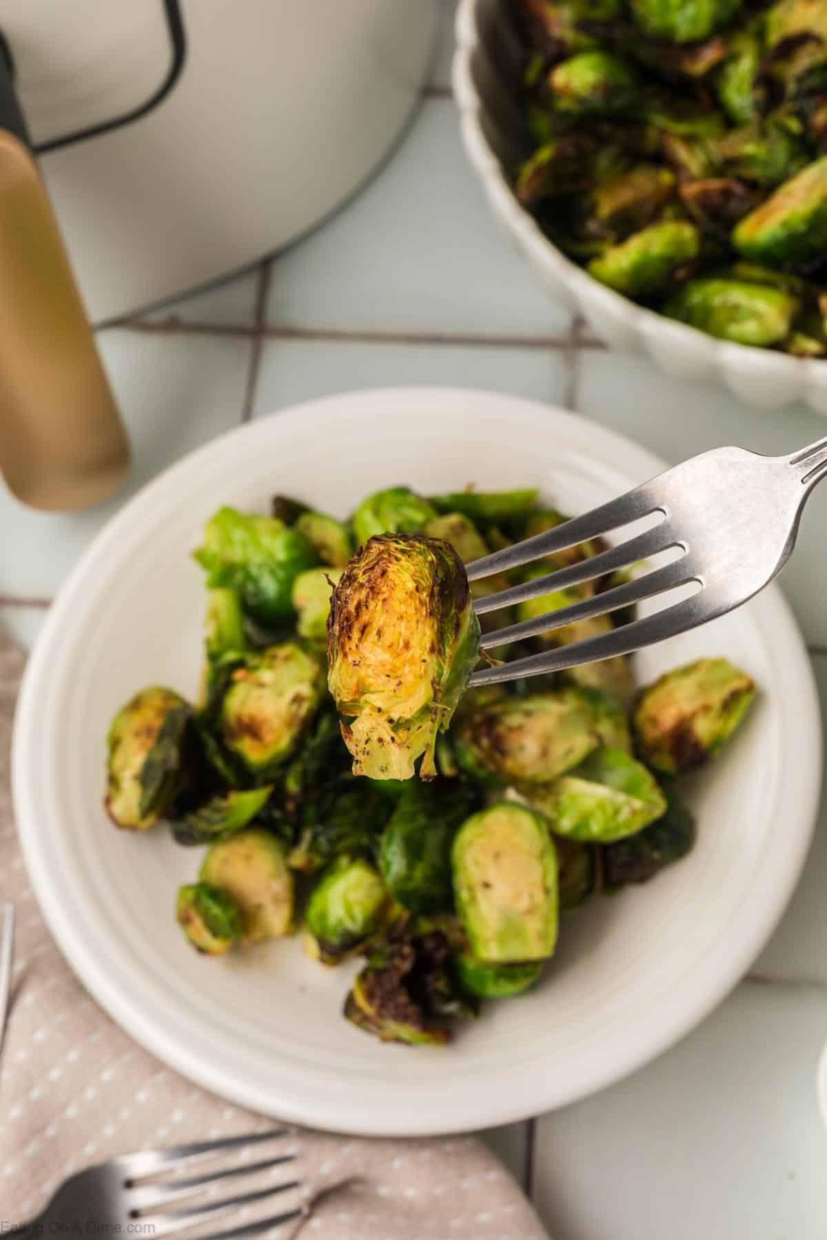 A fork holds a charred Brussels sprout over a white plate filled with roasted Brussels sprouts. A bowl of additional Brussels sprouts and a bottle of dressing are visible in the background, all set on a tiled surface, showcasing an air fryer brussel sprouts recipe at its finest.