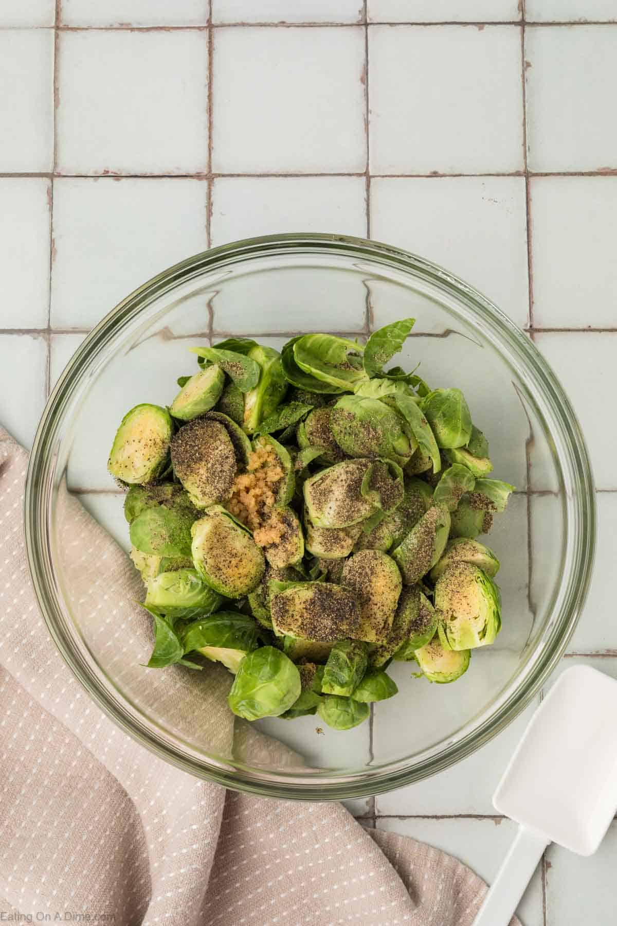 A glass bowl filled with halved Brussels sprouts seasoned with ground black pepper and minced garlic, perfect for an air fryer Brussels sprouts recipe. It's placed on a white tiled surface, accompanied by a white spatula and a beige, textured cloth.