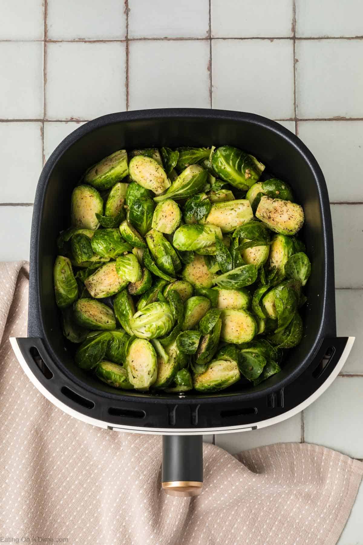 An air fryer basket filled with seasoned Brussels sprouts sits on a tiled countertop, suggesting a delicious air fryer Brussels sprouts recipe. The air fryer rests on a beige cloth with small white polka dots. The Brussels sprouts appear fresh and ready to be roasted.