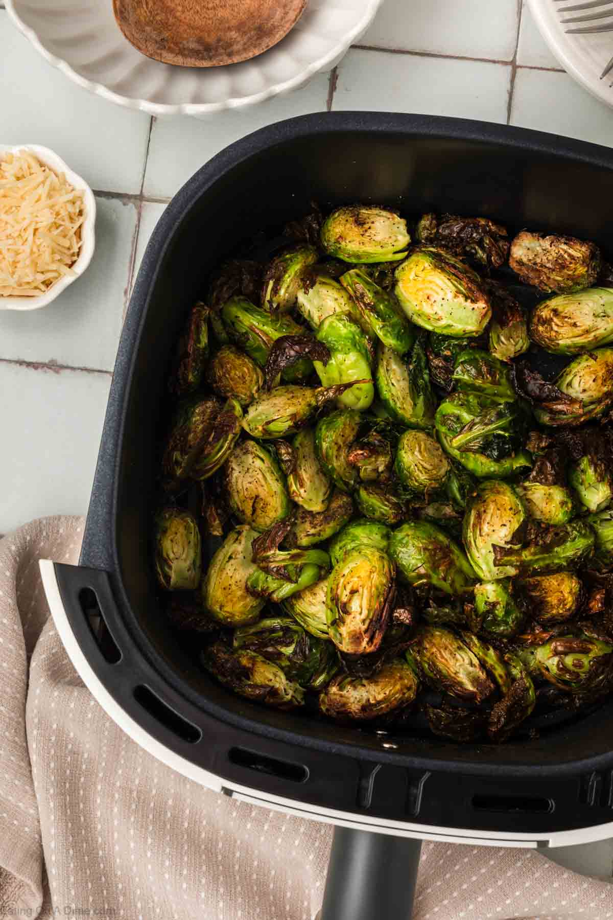 Close-up of roasted Brussels sprouts in an air fryer basket, crispy and golden brown. Surrounding the air fryer are small bowls and a fabric napkin, creating a homey kitchen atmosphere, perfect for trying out your new favorite air fryer Brussels sprouts recipe.