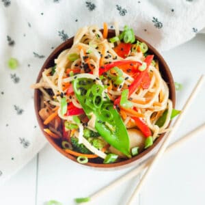 A colorful bowl of Asian noodle salad with sliced red bell peppers, snow peas, shredded carrots, and black sesame seeds sits enticingly next to a patterned napkin. This dish, reminiscent of a vibrant crock pot chicken chow mein recipe, is accompanied by chopsticks resting aside.