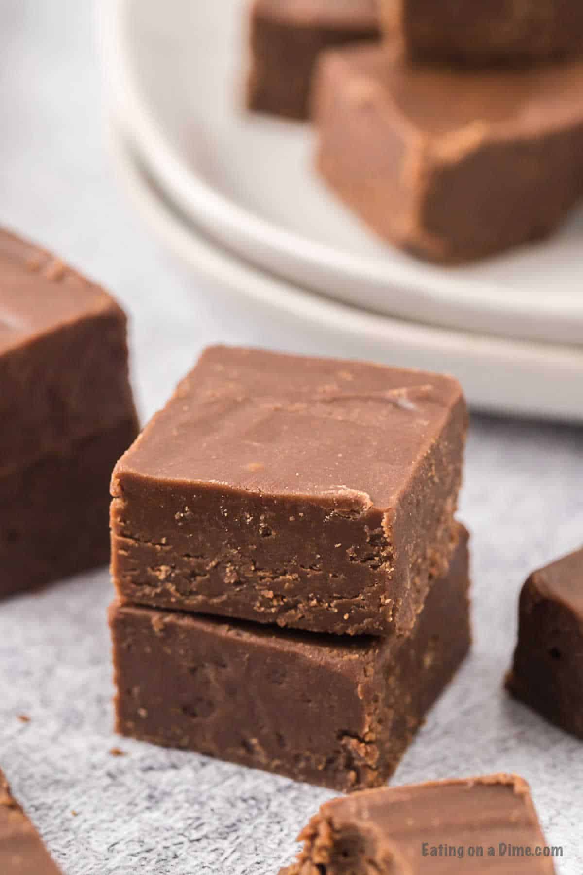 Close-up of homemade 2 Ingredient Fudge cubes stacked on top of each other with a smooth, rich texture. Additional pieces of fudge are visible in the background, with some placed on white plates atop a grey surface.