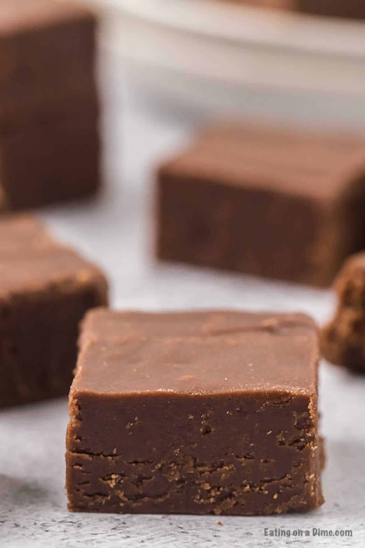 Close-up of several pieces of 2 Ingredient Fudge, with one piece prominently in the foreground. The fudge has a smooth, rich texture and is displayed on a light-colored surface. In the background, more pieces of fudge are slightly blurred.