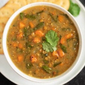 A bowl of curry lentil soup with visible chunks of orange carrots and green vegetables. It's garnished with a sprig of fresh cilantro. In the background, there are round slices of bread on a white plate.