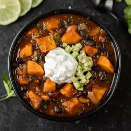 A bowl of crock pot sweet potato black bean chili, brimming with vegetables and topped with diced avocado and a dollop of sour cream. Sliced limes and cilantro garnish the dark background.