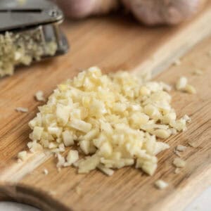 Finely chopped garlic on a wooden cutting board, with a garlic press and unpeeled garlic cloves in the background offers a clear guide on how to mince garlic cloves effectively.