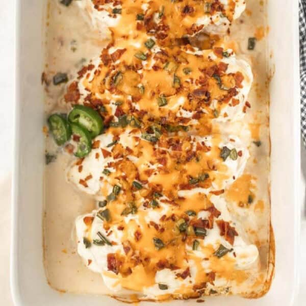 This Keto Jalapeño Popper Chicken dish features baked chicken breasts topped with cream cheese, shredded cheddar, crumbled bacon, and sliced jalapeños in a white casserole dish. The creamy sauce and toppings create a rich and savory appearance perfect for low-carb enthusiasts.