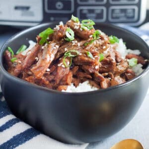 A black bowl brimming with succulent crock pot sesame beef is topped with sesame seeds and chopped green onions over a bed of rice. A striped napkin and a gold spoon rest beside the bowl, with a blurred appliance in the background, highlighting the delicious simplicity of this recipe.