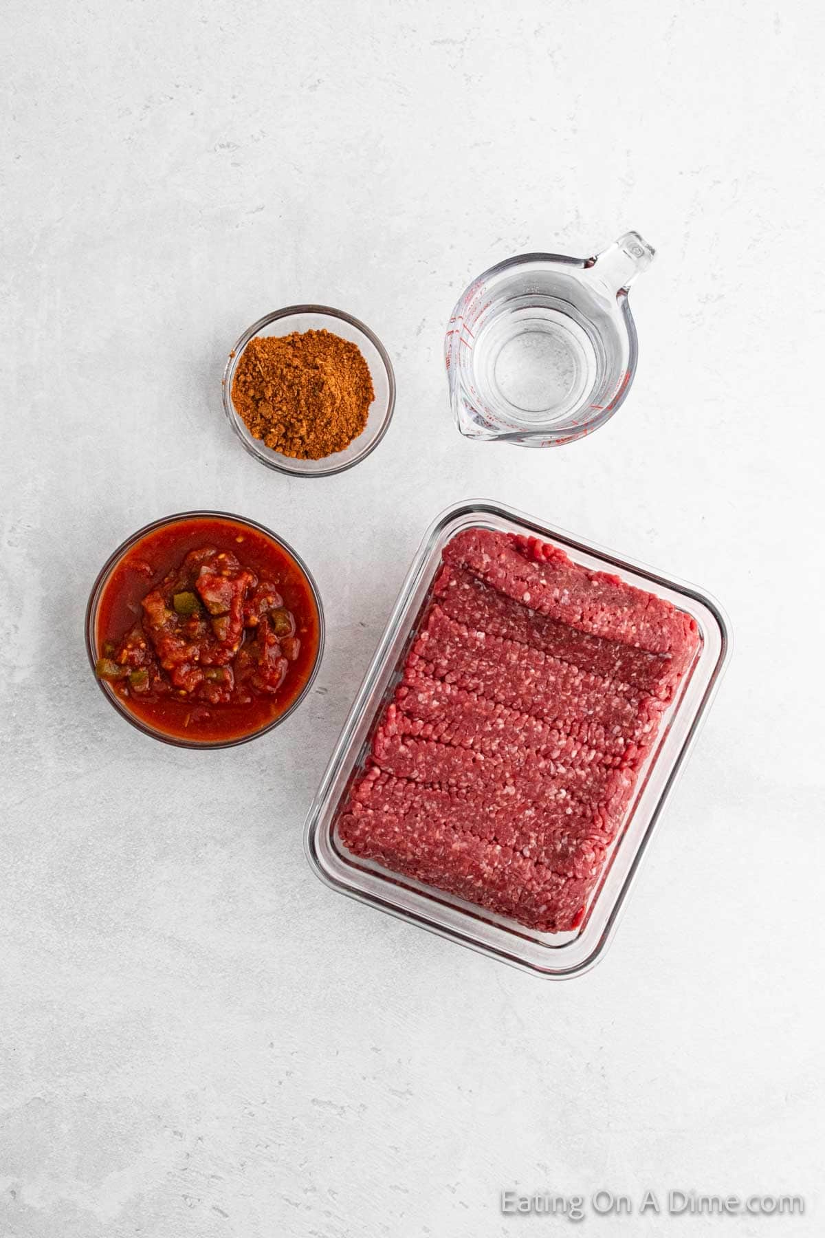 A top-down view of ingredients on a light gray surface, ideal for Instant Pot Taco Meat: a tray of raw minced beef, a bowl of chunky salsa, a small bowl of seasoning, and a measuring cup of water.