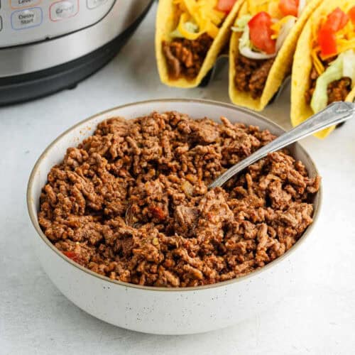 A bowl of cooked ground beef with a spoon inside sits on a table, showcasing delicious Instant Pot taco meat. In the background, hard shell tacos are filled with beef, shredded cheese, lettuce, and diced tomatoes. An Instant Pot is partially visible on the side.