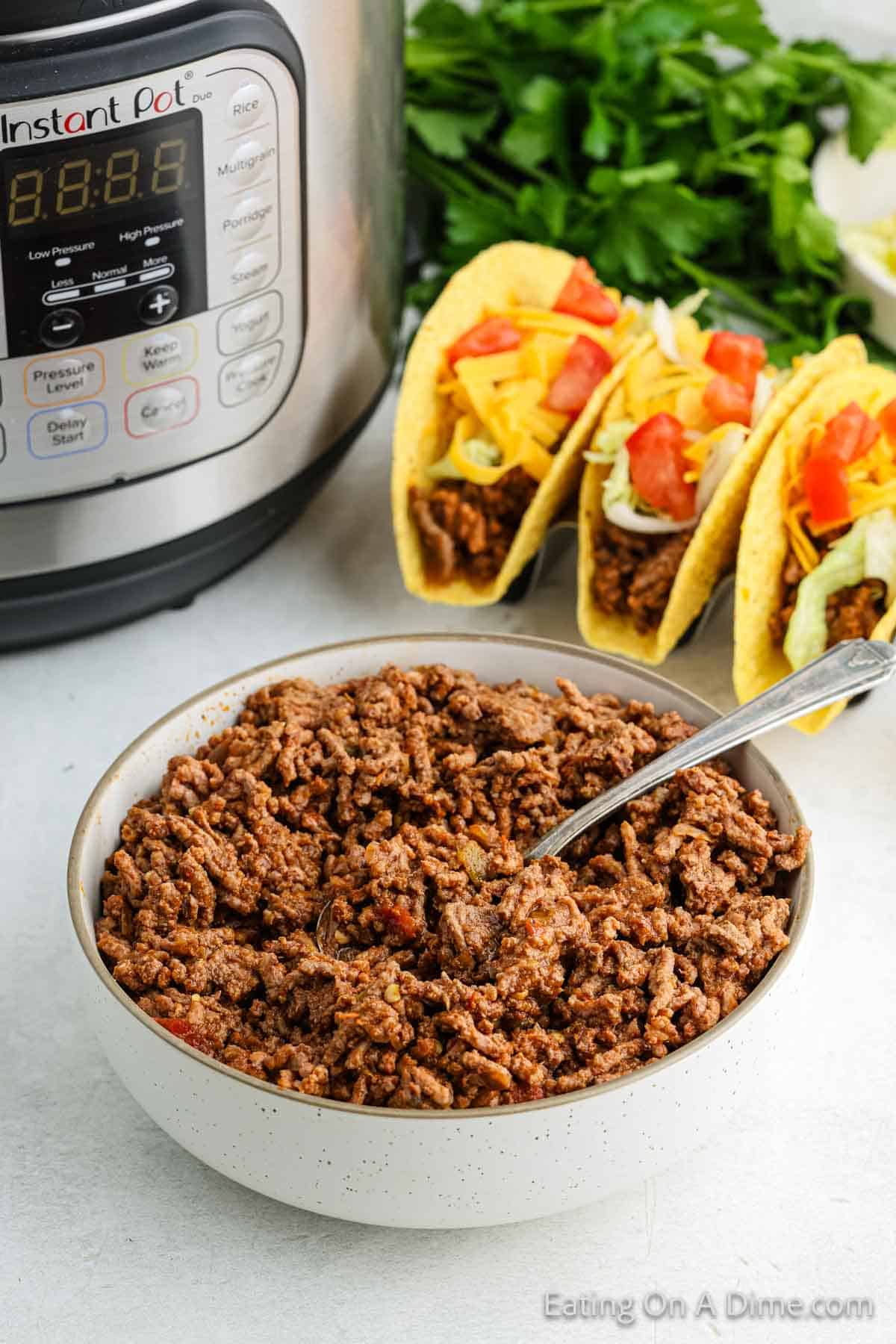 A bowl of Instant Pot taco meat sits in front of the cooker. Three hard-shell tacos filled with this savory ground beef, crisp lettuce, cheese, and tomatoes are in the background, alongside fresh parsley on a light-colored surface.