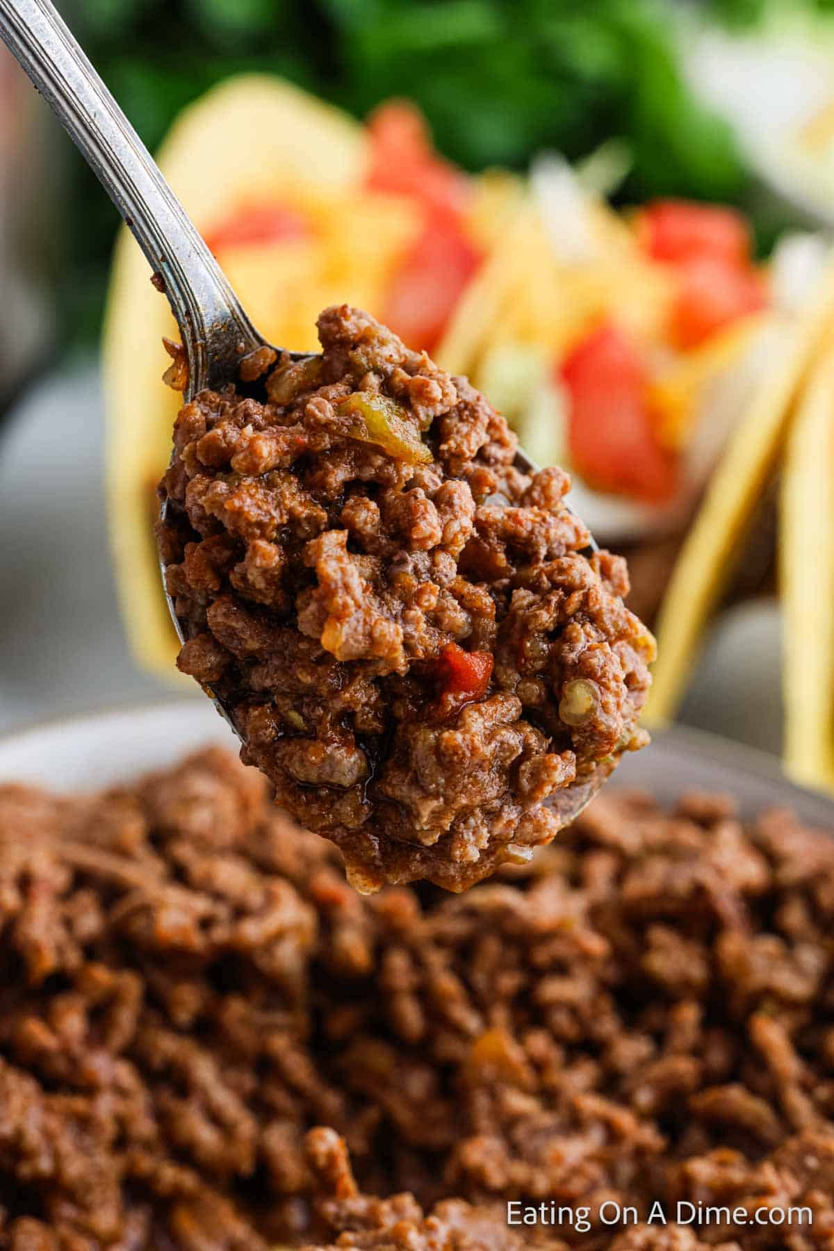 Close-up of a spoonful of seasoned Instant Pot taco meat mixed with diced vegetables. In the background, hard shell tacos brim with colorful toppings like tomatoes and lettuce, suggesting a vibrant and appetizing meal preparation.