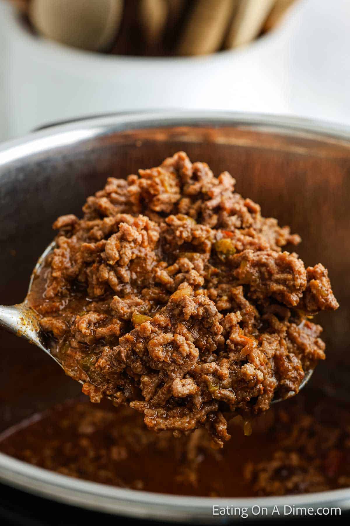 A spoonful of Instant Pot taco meat with spices is held over a pot, showcasing its texture and savory appearance. The background features a blurred container with kitchen utensils, highlighting the perfect blend of convenience and flavor in this dish.