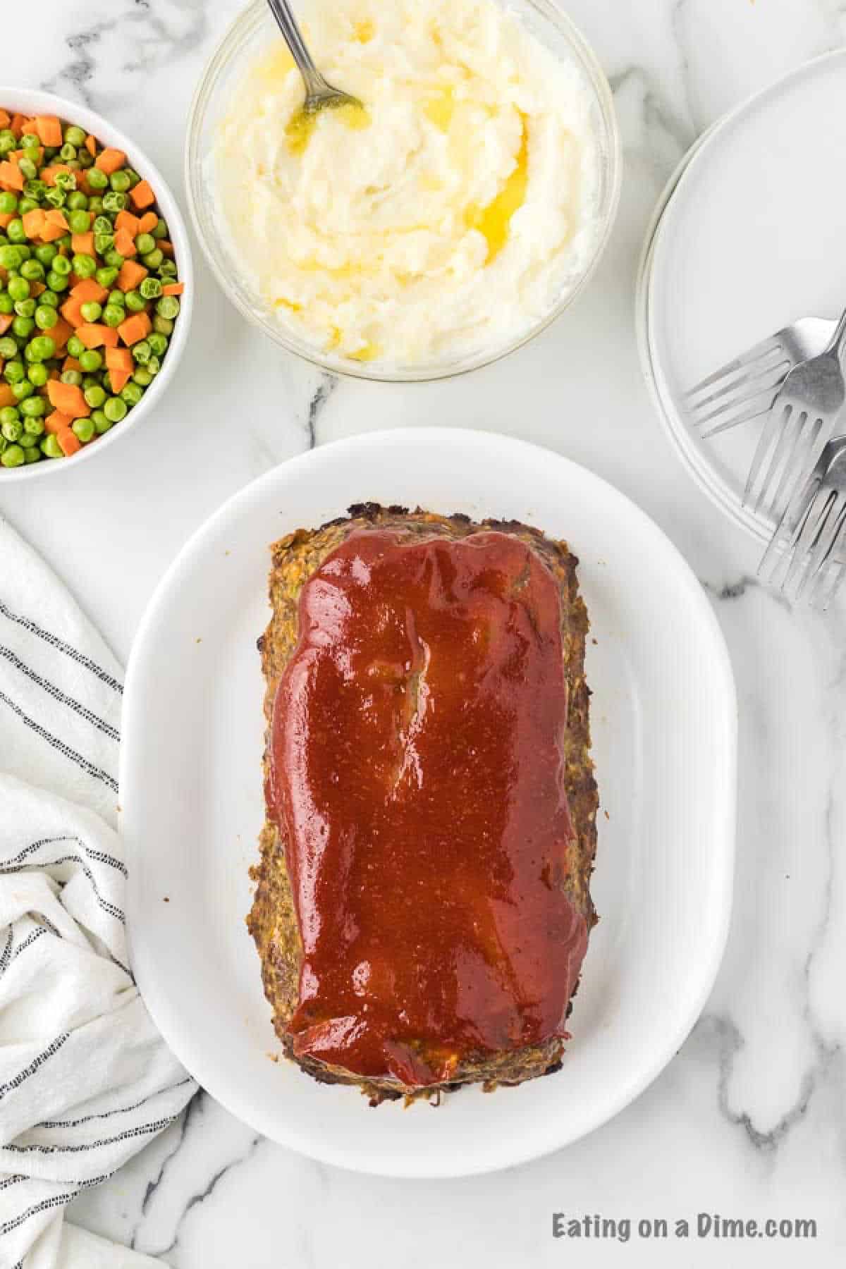 Close up image of meatloaf on a white plate with a side of mixed vegetables and mashed potatoes. 