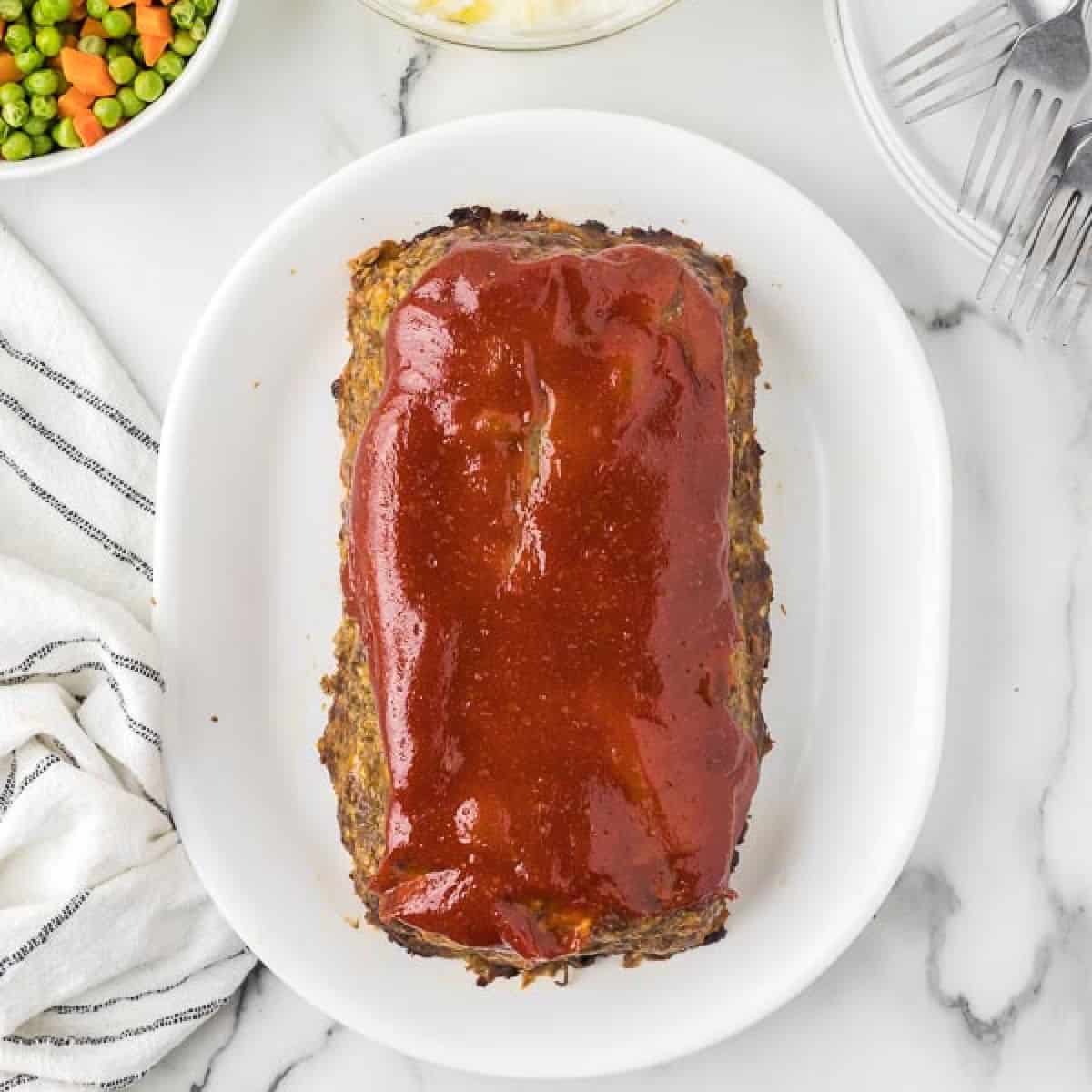 A glazed meatloaf, reminiscent of a classic Cracker Barrel recipe, sits on a white oval plate on a marble surface. Nearby are forks, a striped cloth, and a bowl of mixed peas and carrots.